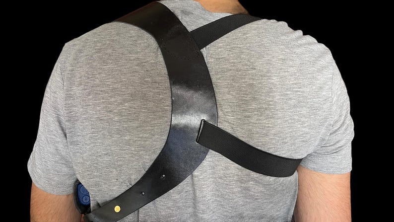 May include: Black leather shoulder holster with a cross-body design. The holster is worn over a gray t-shirt. The holster has a strap that goes over the shoulder and another that goes around the torso. A gold-colored rivet is visible.