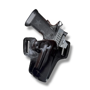 Peut inclure: Un pistolet noir avec une étoile sur la poignée est partiellement inséré dans un étui en cuir noir. L'étui est cousu et a une finition brillante. L'arme a un composant de couleur argent.