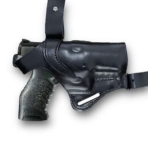 May include: Black leather shoulder holster with a semi-automatic pistol. The holster has a molded design to fit the gun and includes adjustable straps for a secure fit. The pistol grip is textured for a firm hold.