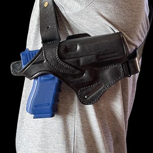 May include: Black leather shoulder holster with a blue handgun. The holster is worn over a gray shirt. The holster has a strap that goes over the shoulder and around the back. The handgun is visible in the holster.