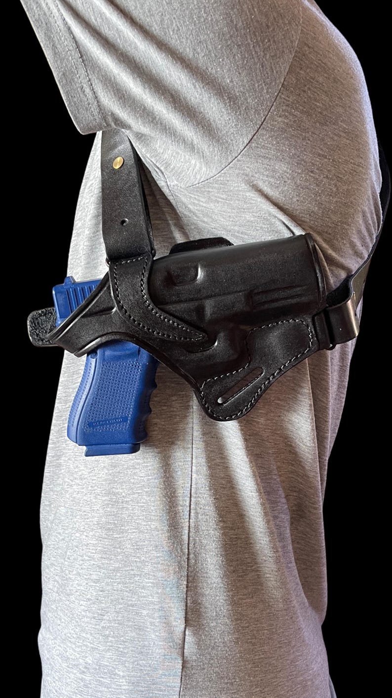 May include: Black leather shoulder holster with a blue handgun. The holster is worn over a gray shirt. The holster has a strap that goes over the shoulder and around the back. The handgun is visible in the holster.