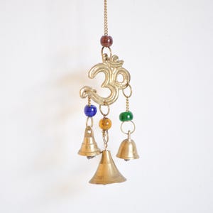 May include: A brass-colored wind chime featuring the Om symbol, with three small bells hanging below. The chime has colorful beads in red, blue, yellow, and green. The bells are suspended from a chain.