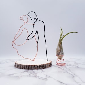 May include: A wire sculpture of a couple dancing, made with copper and black wire. The sculpture is on a wooden base and next to a small air plant in a copper spring.