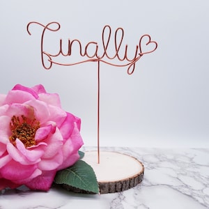 May include: A copper wire sculpture of the word "Finally" with a heart at the end, displayed on a wooden slice with a pink rose.