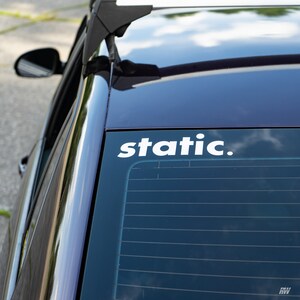 May include: A white vinyl decal applied to the rear window of a dark-coloured vehicle. The decal features the word static. in a lowercase, sans-serif font. The car has a black roof rack mount visible at the top edge of the window frame.