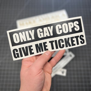 May include: A black and white bumper sticker with the text "ONLY GAY COPS GIVE ME TICKETS" in a bold, sans-serif font. The sticker is rectangular and held in a hand, with other similar stickers visible in the background. The background is a dark gray surface.