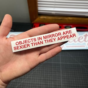 May include: A white rectangular sticker with the text "OBJECTS IN MIRROR ARE SEXIER THAN THEY APPEAR" in red. The sticker is held in a hand.