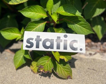 May include: A rectangular white sticker featuring the word static. printed in a bold, black, sans-serif font. The sticker is positioned horizontally, displaying the lowercase letters s, t, a, t, i, c, followed by a period at the end.