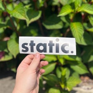 May include: A rectangular white sticker featuring the word static. printed in a bold, black, sans-serif font. The sticker has a clean, minimalist design with a full stop at the end of the text, presented on a clear backing material.