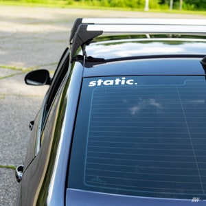 May include: A white vinyl decal with the word static. in a lowercase sans-serif font applied to the upper corner of a dark-coloured car's rear window. The vehicle features a black roof rack system mounted above the tinted glass rear windscreen.