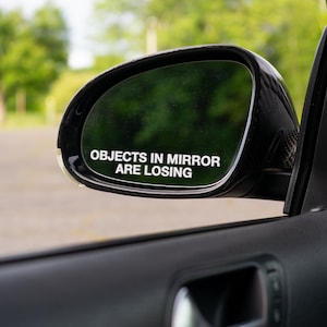 May include: A black car side mirror with the text "OBJECTS IN MIRROR ARE LOSING" in white. The mirror reflects a blurred green background. The car door and handle are visible in the foreground.