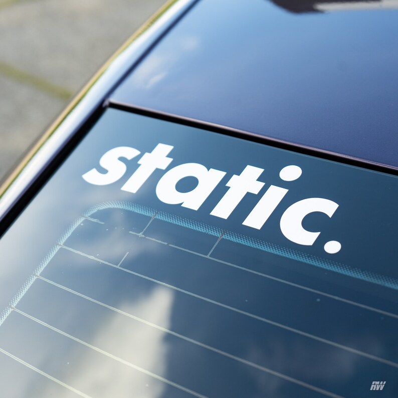 May include: A white vinyl decal applied to the rear window of a dark-coloured vehicle. The decal features the word static in a bold, lowercase sans-serif font, followed by a period. The text is positioned horizontally across the upper glass.