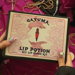 Puede incluir: Una caja negra con una ilustración rosa y blanca de dos manos sosteniendo una daga roja. La caja dice "Gayuma Lip Potion DIY Lip Gloss Kit" y "Love" en la parte superior e inferior. La caja está rodeada de cadenas doradas, collares de perlas y velas de té.
