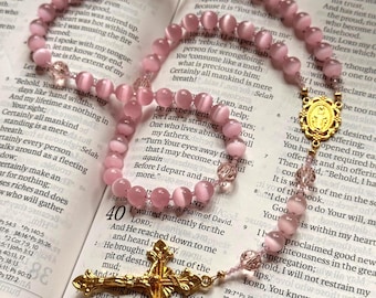 May include: A rosary featuring smooth, light pink cat's eye beads, clear faceted accent beads, a gold-toned metal centerpiece depicting a religious figure, and a detailed gold-toned metal crucifix, all resting on the open pages of a printed book.