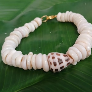 May include: A bracelet made of white seashells, with a gold-coloured clasp. The bracelet features small, round, white shells and a larger, patterned shell in the centre. It rests on a green leaf.