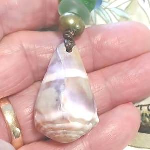 May include: A handcrafted necklace featuring a teardrop-shaped seashell pendant with purple and white hues. The pendant is accented with blue and green sea glass beads, a pearl, and a brown cord. This unique jewelry piece is perfect for beach lovers.