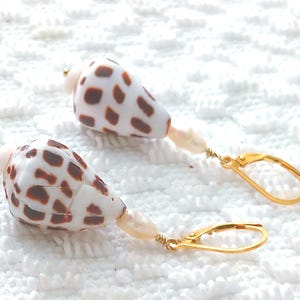 May include: A pair of seashell earrings. Each earring features a white shell with brown spots, a small pearl, and a gold-colored leverback clasp. The earrings are displayed on a white, textured surface.