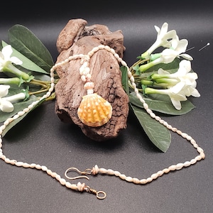 Op de afbeelding: Een witte schelpketting met een grotere schelp hanger. De hanger is licht oranje van kleur met bruine markeringen. De ketting ligt op een houten oppervlak met witte bloemen op de achtergrond. Er zijn ook bijpassende oorbellen.