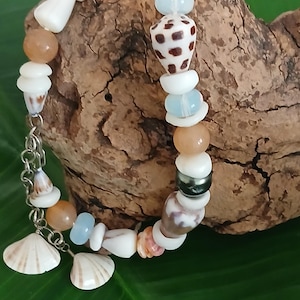 Puede incluir: Una pulsera de cuentas con una variedad de conchas marinas naturales, perlas y cuentas de colores. La pulsera incluye cuentas blancas, marrones y azules, con una cadena plateada y dijes de conchas marinas. La pulsera se exhibe sobre un trozo de madera flotante.