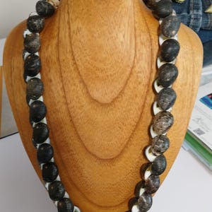 May include: A handcrafted necklace displayed on a wooden bust. The necklace features alternating dark, speckled beads and white, shell-shaped pieces. The beads have a natural, organic appearance, creating a unique accessory.
