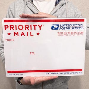 May include: A white Priority Mail shipping box with red accents and text. The box features the USPS logo and the words "PRIORITY MAIL" and "UNITED STATES POSTAL SERVICE." The box is held by a person.
