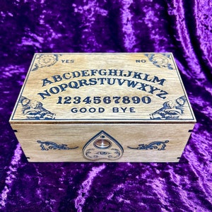 May include: A wooden Ouija board with a planchette in the shape of a teardrop. The board has the letters of the alphabet, numbers 1-9 and 0, the words "yes", "no", and "goodbye", and two images of spirits. The board is on a purple velvet surface.