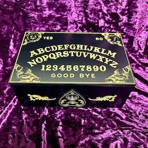 Puede incluir: Una tabla de Ouija de madera negra con letras doradas y una planchette. La tabla presenta el alfabeto, los números del 1 al 9 y el 0, las palabras "sí", "no" y "adiós".