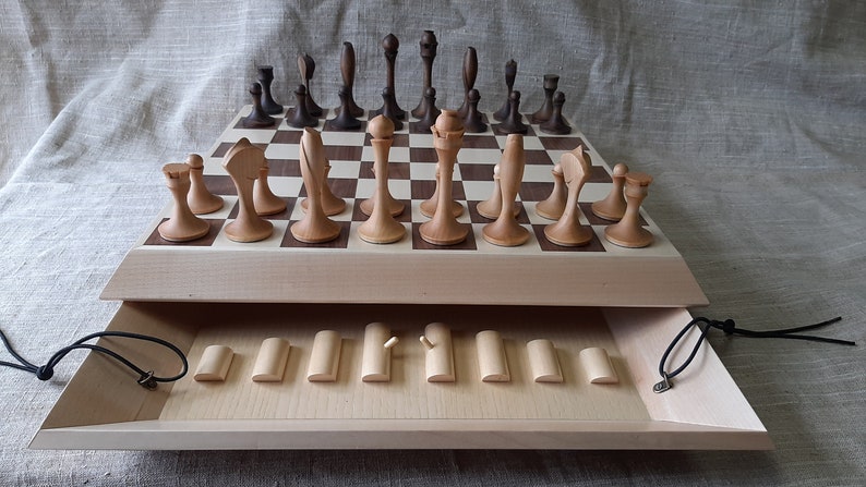 Pode incluir: Um jogo de xadrez de madeira com um tabuleiro de madeira clara e pe&ccedil;as de madeira escura. O tabuleiro tem uma tampa com dobradi&ccedil;as que se abre para revelar um compartimento para guardar as pe&ccedil;as. As pe&ccedil;as s&atilde;o de madeira e t&ecirc;m um acabamento natural.