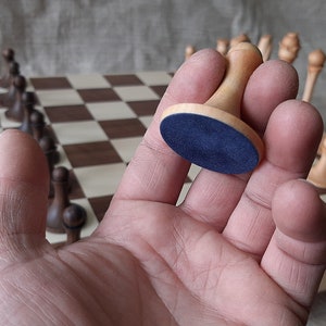 Pode incluir: Uma pe&ccedil;a de xadrez de madeira com uma base de feltro azul. A pe&ccedil;a est&aacute; sendo segurada em uma m&atilde;o sobre um tabuleiro de xadrez com outras pe&ccedil;as de xadrez.