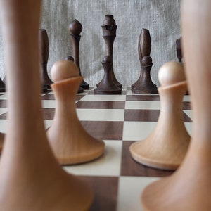 Pode incluir: Uma vis&atilde;o de perto de um tabuleiro de xadrez de madeira com quadrados claros e escuros. O tabuleiro est&aacute; configurado com pe&ccedil;as de xadrez, incluindo pe&otilde;es, torres e um rei. As pe&ccedil;as s&atilde;o feitas de madeira e t&ecirc;m um acabamento natural.