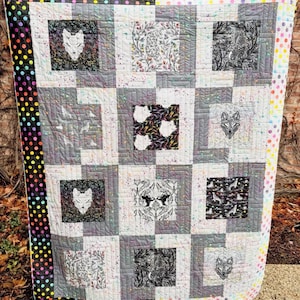 May include: A handmade quilt with a patchwork design. The quilt features square blocks with various patterns, including wolf faces, floral designs, and animal silhouettes. The border is a mix of polka dots and solid colors. The color palette includes gray, white, black, and splashes of color.