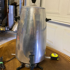 May include: A vintage, stainless steel coffee percolator with a black handle and lid finial. The coffee maker has a spigot and sits on a black base with three legs. A black power cord is attached. The percolator is on a wooden surface.