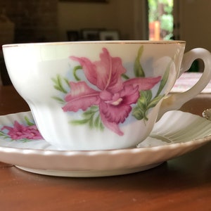 May include: A white teacup and saucer with a pink orchid floral design. The cup and saucer have a gold trim.