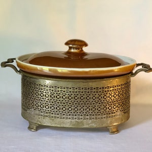 Puede incluir: Una fuente de cerámica marrón con tapa y un soporte de metal decorativo con diseño de celosía. El soporte tiene tres patas y es de forma ovalada.