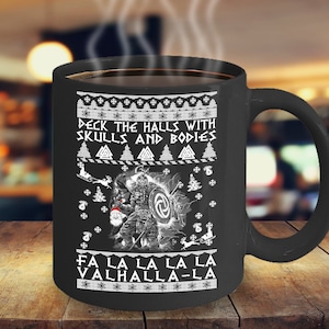 May include: Black coffee mug with a white design featuring a Viking warrior holding a shield and a Christmas tree. The text reads "Deck the halls with skulls and bodies" and "Fa la la la la Valhalla-la".