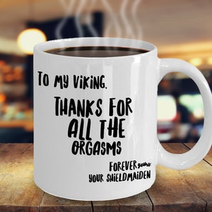 May include: White ceramic mug with a curved handle, filled with a dark liquid. The mug features black text that reads "To my Viking, Thanks for all the orgasms, Forever yours, Your Shieldmaiden."