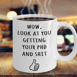May include: White ceramic coffee mug with black text that reads "WOW, LOOK AT YOU GETTING YOUR PHD AND SHIT" with a thumbs up emoji at the bottom.