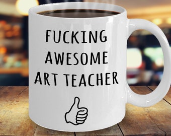 May include: White ceramic coffee mug with a black inscription that reads "FUCKING AWESOME ART TEACHER" with a thumbs up icon below the text.