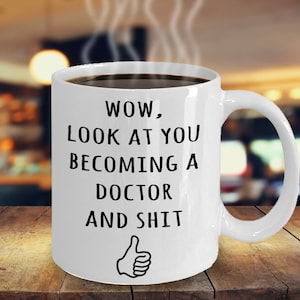 May include: White coffee mug with black text that reads "WOW, LOOK AT YOU BECOMING A DOCTOR AND SHIT" with a thumbs up emoji.