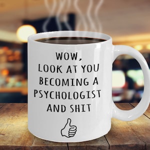 May include: A white ceramic mug filled with dark liquid, possibly coffee or tea, with steam rising. The mug has black text that reads "WOW, LOOK AT YOU BECOMING A PSYCHOLOGIST AND SHIT" with a thumbs-up graphic. The mug is on a wooden surface.