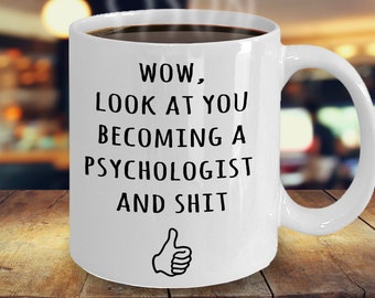 May include: A white ceramic mug filled with dark liquid, possibly coffee or tea, with steam rising. The mug has black text that reads "WOW, LOOK AT YOU BECOMING A PSYCHOLOGIST AND SHIT" with a thumbs-up graphic. The mug is on a wooden surface.