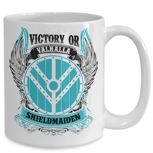 Könnte beinhalten: Weiße Keramik-Kaffeetasse mit einem schwarzen und blauen Design. Das Design zeigt einen Schild mit einer blauen Mitte und schwarzem Rand, Flügeln und dem Text "Victory or Valhalla Shieldmaiden".