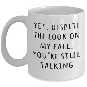 May include: White ceramic coffee mug with black text that reads "Yet, despite the look on my face, you're still talking".