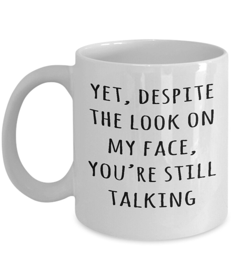 May include: White ceramic coffee mug with black text that reads "Yet, despite the look on my face, you're still talking".