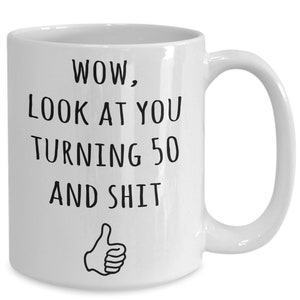 May include: White ceramic coffee mug with black text that reads "WOW, LOOK AT YOU TURNING 50 AND SHIT" with a thumbs up icon.