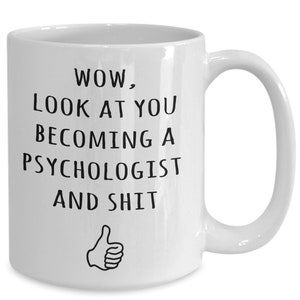 May include: White ceramic mug with a black text that reads "WOW, LOOK AT YOU BECOMING A PSYCHOLOGIST AND SHIT" with a thumbs-up graphic. The mug has a curved handle and is designed for hot beverages.