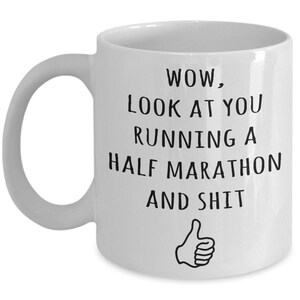 May include: White ceramic coffee mug with black text that reads "WOW, LOOK AT YOU RUNNING A HALF MARATHON AND SHIT" with a thumbs up emoji.