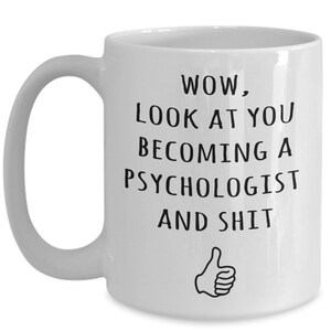 May include: White ceramic mug with a black text that reads "WOW, LOOK AT YOU BECOMING A PSYCHOLOGIST AND SHIT" with a thumbs-up graphic. The mug has a curved handle.