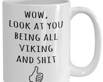 Puede incluir: Taza de cerámica blanca con texto negro: "WOW, MIRA CÓMO ERES TODO VIKINGO Y MIERDA" y un gráfico de pulgar hacia arriba. La taza tiene un asa curva y es adecuada para bebidas frías o calientes.