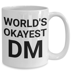 May include: White ceramic coffee mug with black text that reads "WORLD'S OKAYEST DM".